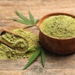 Bowl of hemp protein with two small hemp leaves and a wooden spoon on a wooden table, showcasing natural, plant-based nutrition